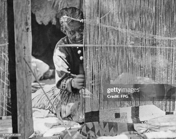 An elderly Native American Navajo woman, dressed in traditional Navajo clothing, weaving a rug on a Navajo reservation in northwestern New Mexico,...