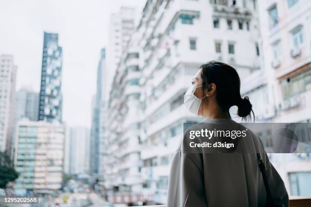 young asian woman with protective face mask to protect and prevent from the spread of viruses in the city, standing on the urban bridge, overlooking the city skyline. in hope that the pandemic will end soon - corona landmarks stockfoto's en -beelden