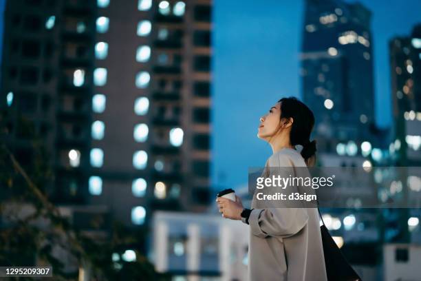 thoughtful young asian woman having a cup of coffee, looking up to sky while standing against highrise illuminated city buildings in the evening - looking to the future photos et images de collection