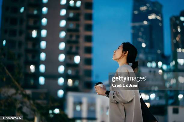 thoughtful young asian woman having a cup of coffee, looking up to sky while standing against highrise illuminated city buildings in the evening - asian woman dream stockfoto's en -beelden