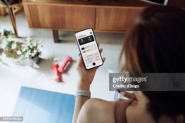 over the shoulder view of young asan sports woman refreshing with water while managing online banking on mobile app with smartphone after fitness work out at home. transferring money, paying bills, checking balances. technology makes life so much easier - hi tech sports gear foto e immagini stock