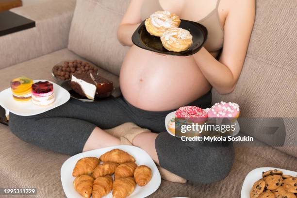 Close up of hungry pregnant woman sitting on the sofa is eating a lot of unhealthy food such as donuts, cakes, chocolate cereal balls, croissants and cookies. Sweet cravings during pregnancy concept