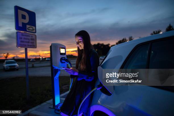 vrouw die haar elektrische auto op benzinepost bij nacht laadt. vrouw die mobiele telefoon gebruikt terwijl het wachten op elektrische auto om in de parkerenpartij bij nacht op te laden - electric people stockfoto's en -beelden