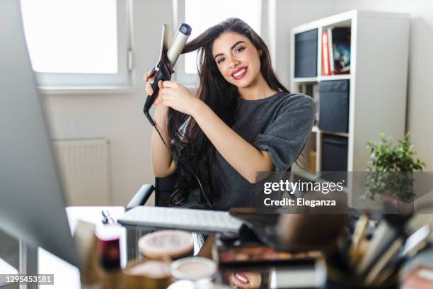 woman looking at computer and using her curling iron while sitting at the dressing table. woman does her hair by watching a tutorial on the internet at the time of the corona virus pandemic. - hair curlers stock pictures, royalty-free photos & images