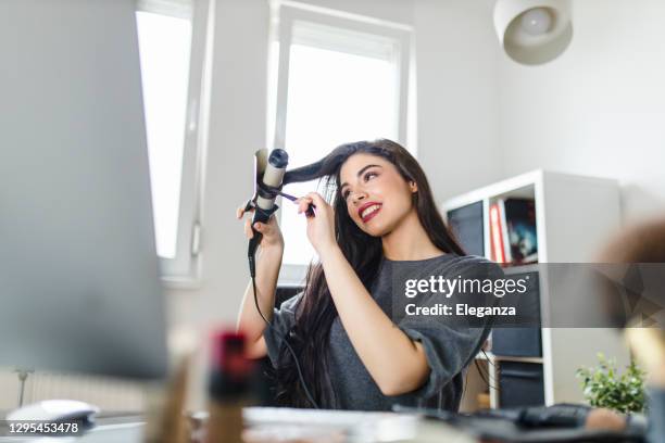 frau schaut auf computer und mit ihrem lockenbügeleisen, während sie am schminktisch sitzt. frau macht ihre haare, indem sie ein tutorial im internet zum zeitpunkt der corona-virus-pandemie. - lockenstab stock-fotos und bilder