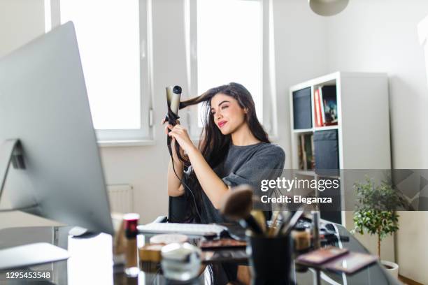 frau schaut auf computer und mit ihrem lockenbügeleisen, während sie am schminktisch sitzt. frau macht ihre haare, indem sie ein tutorial im internet zum zeitpunkt der corona-virus-pandemie. - lockenstab stock-fotos und bilder