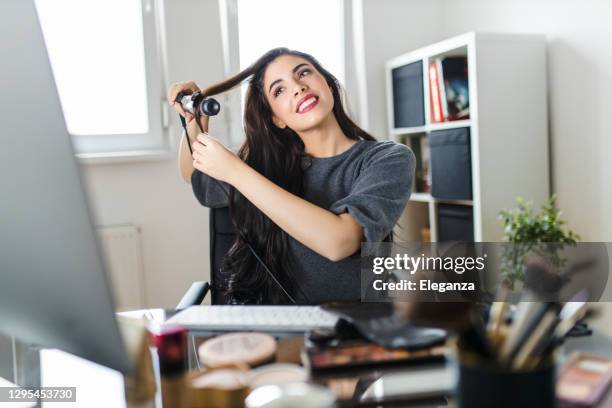 mulher olhando para o computador e usando seu ferro de enrolar enquanto se senta na penteadeira. mulher faz seu cabelo assistindo um tutorial na internet no momento da pandemia do vírus corona. - curled up - fotografias e filmes do acervo