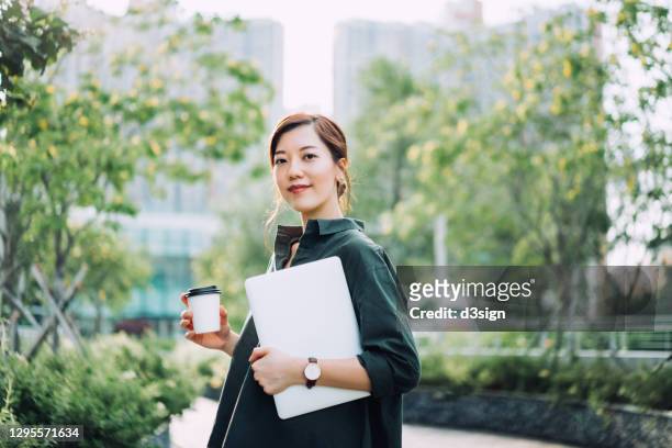 portrait of confident and successful young asian businesswoman holding laptop with a cup of coffee, standing in urban office park against green nature plants. business on the go - asiático e indiano imagens e fotografias de stock
