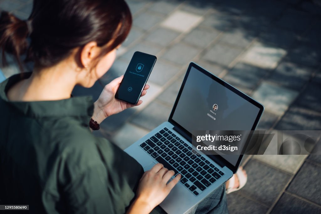 Young Asian businesswoman sitting on the bench in an urban park working outdoors, logging in to her laptop and holding smartphone on hand with a security key lock icon on the screen. Privacy protection, internet and mobile security concept
