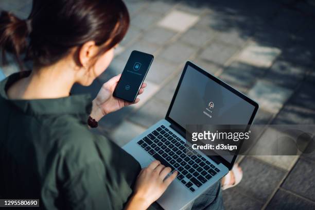 young asian businesswoman sitting on the bench in an urban park working outdoors, logging in to her laptop and holding smartphone on hand with a security key lock icon on the screen. privacy protection, internet and mobile security concept - protezione foto e immagini stock