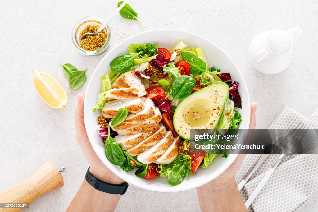 Gegrilltes Hühnerfleisch und frischer Gemüsesalat aus Tomaten, Avocado, Salat und Spinat. Gesundes und entgiftendes Lebensmittelkonzept. Ketogene Ernährung. Buddha Schale in den Händen auf weißem Hintergrund, Ansicht von oben