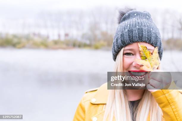 blonde woman with red lips smiling and a grey wool cap covering her left eye with a yellow leaf. water bottom of the lake and trees without leaves. - lovely frozen leaves stock pictures, royalty-free photos & images