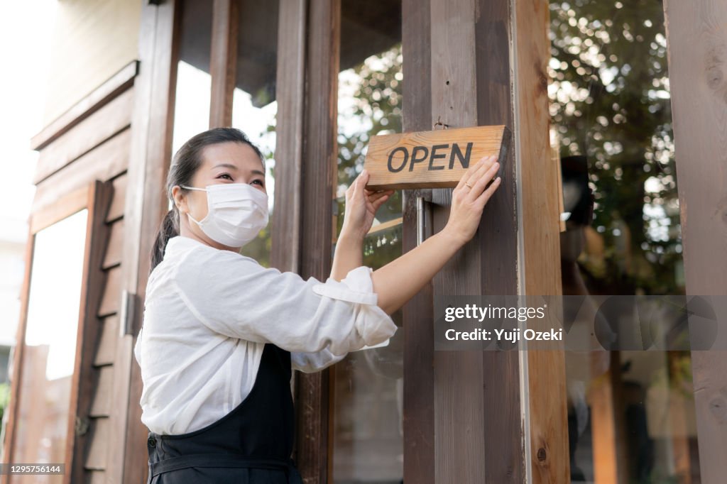 Portrait shot of restaurant manager with face mask ,holding an open sign at the entrance as preparation to reopening post pandemic Covid 19