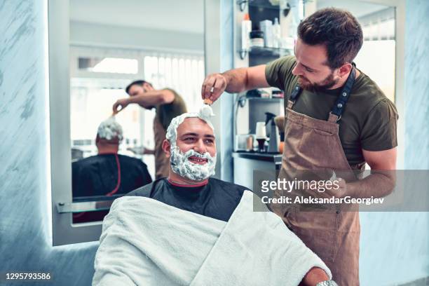 het mannelijke glimlachen terwijl het krijgen voorbereid voor baard en hoofd scheert door kapper - shaving cream stockfoto's en -beelden