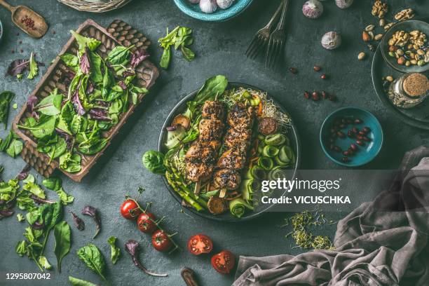bowl with fried meat skewers with green salad of cucumber, asparagus and lettuce on dark kitchen table background with ingredients and forks. - paleo imagens e fotografias de stock