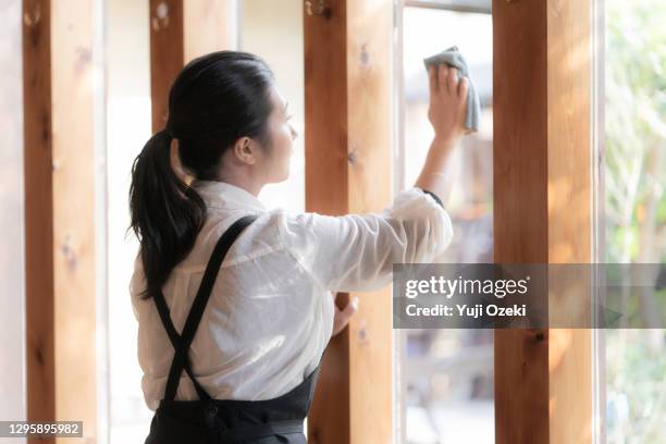 asian young woman wiping the window glass of a cafe restaurant wearing an apron with a ponytail - deeltijdbaan stockfoto's en -beelden