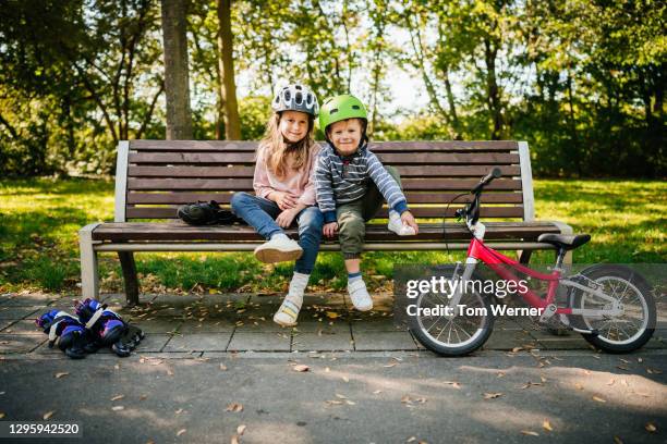 portrait of two young siblings wearing bike helmets sitting on park bench - schuhe für sport und freizeit stock-fotos und bilder