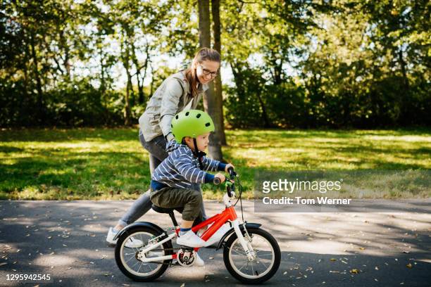 mom guiding young son learning to ride bike in the park - radfahren stock-fotos und bilder