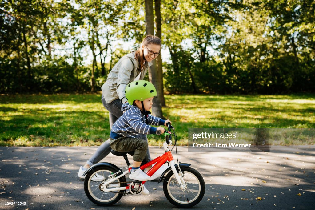 Mom Guiding Young Son Learning To Ride Bike In The Park