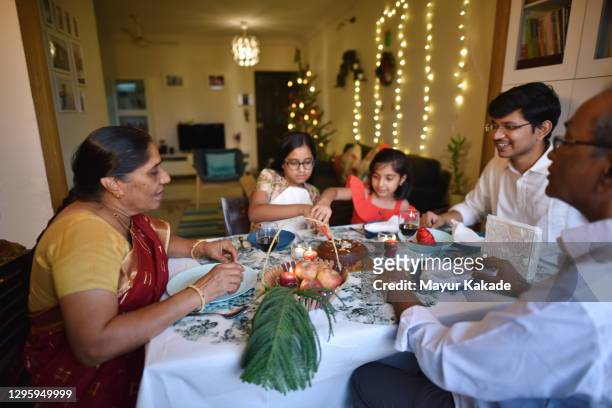 two girls cutting cake with family on a dining table on christmas eve in a house decorated with christmas tree and string lights - indian family dinner table stock pictures, royalty-free photos & images