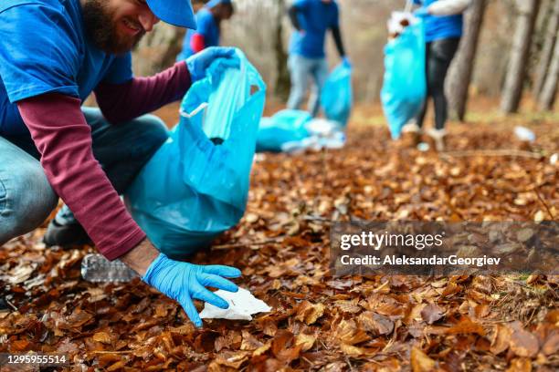 male volunteer squatting to pick up trash from forest grounds and put it in bag - macedonia country stock pictures, royalty-free photos & images