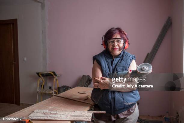 mature woman bricklayer poses with radial saw looking at camera in the remodeling of a room of a house, ear and eye protection - female bricklayer stock pictures, royalty-free photos & images