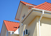A stucco house with a red metal roof, attic and air conditioner outdoor unit with a close-up on a white roof soffit board and rain gutter system with a downspout against blue sky.