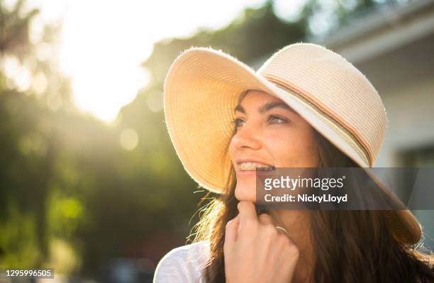 晴れた午後に外に立っている太陽の帽子をかぶった笑顔の若い女性 - adult retainer ストックフォトと画像