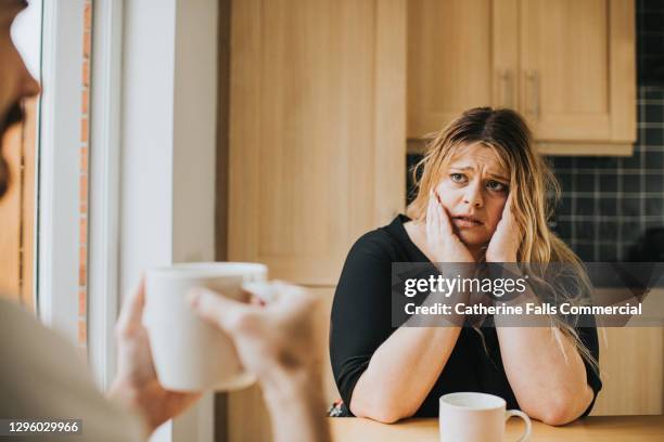 worried woman sits at a kitchen table holding her head in her hands - cross stockfoto's en -beelden