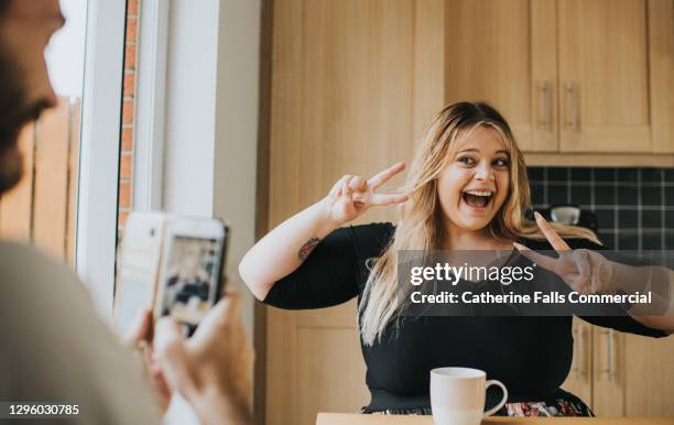 confident woman makes a v-sign with her hands as a man takes a photo on a phone - two lovers photocall stock-fotos und bilder