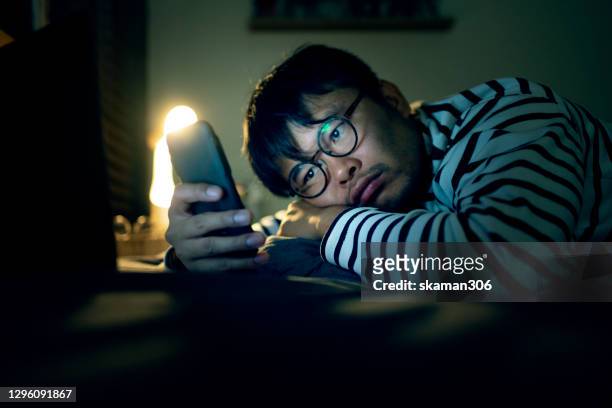 asian young adult man feeling stressed and boring using mobile phone and laptop on bed at night times - digital detox stock pictures, royalty-free photos & images