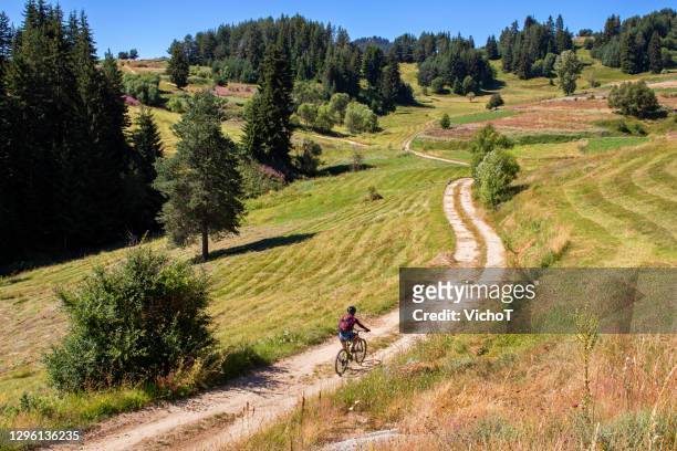young woman riding bike along dirt road surrounded by grass fields and flowers in a mountain area - bulgaria stock pictures, royalty-free photos & images