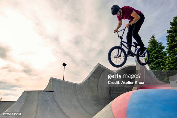 male rider with bmx bike jumping on concrete ramp against cloudy sky at skateboard park - bmx stock-fotos und bilder