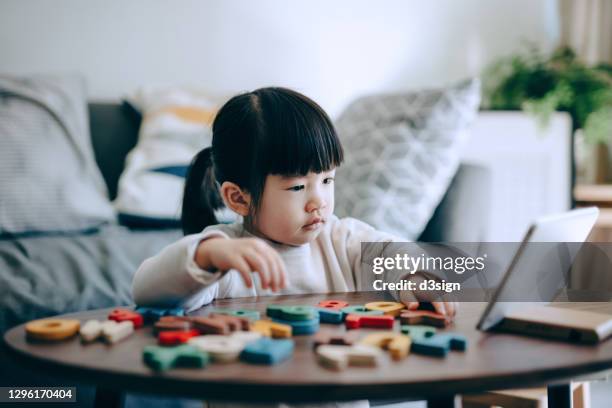 lovely little asian girl studying online with digital tablet at home. she is learning alphabets and playing with colourful wooden capital letters. family lifestyle, homeschooling and technology theme - spelling stockfoto's en -beelden