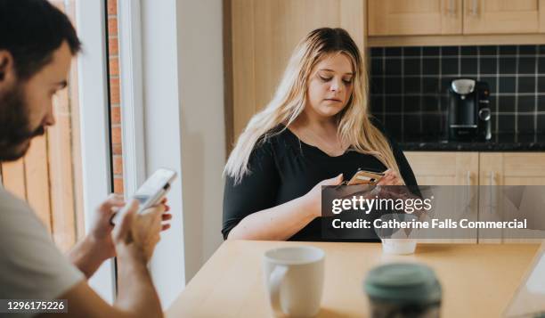 a couple ignore each other at the kitchen table, while they both stare down at their mobile phones - negatieve eigenwaarde stockfoto's en -beelden