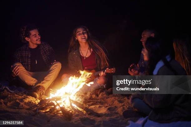 gruppo di amici che cantano suonando ukulele al falò notturno in spiaggia - bon fire foto e immagini stock