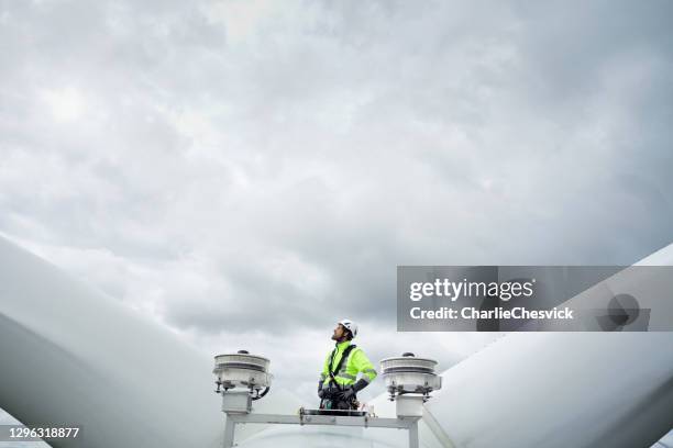 técnico profesional de acceso a cuerdas de pie en el techo (hub) de turbina de viento entre las cuchillas y las antenas y mirando hacia arriba en la hoja. cielo dramático detrás. manos en las caderas - aerogenerador fotografías e imágenes de stock