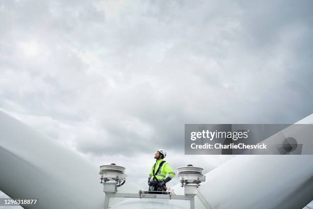 técnico profesional de acceso a cuerdas de pie en el techo (hub) de turbina de viento entre las cuchillas y las antenas y mirando hacia arriba en la hoja. cielo dramático detrás. manos en las caderas - servizio fotografico fotografías e imágenes de stock