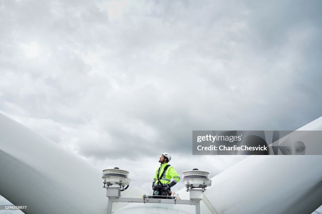 Professional rope access technician standing on roof (hub) of wind turbine between blades and antennas and looking up on the blade. Dramatic sky behind. Hands on hips