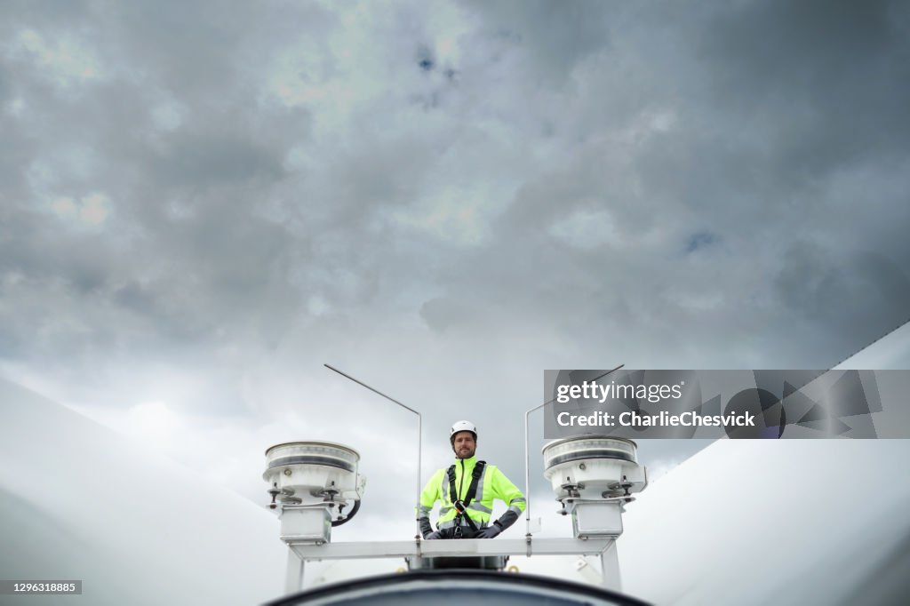 Professionell repåtkomsttekniker som står på tak (nav) på vindkraftverk mellan blad och antenner och ser till kameran. Dramatisk himmel bakom. Händerna på höfterna