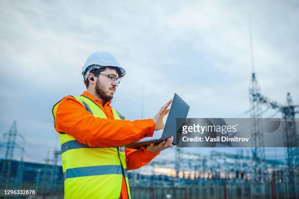 ingeniero eléctrico trabajando en un ordenador portátil frente a la foto de la estación de energía eléctrica - suministro de energía fotografías e imágenes de stock
