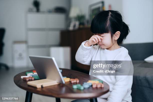 exhausted little asian girl rubbing tired eyes while studying online from home. she is attending online school classes with a digital tablet, with colourful letters learning materials on coffee table. family lifestyle, homeschooling and technology theme - encouragement letter stock pictures, royalty-free photos & images