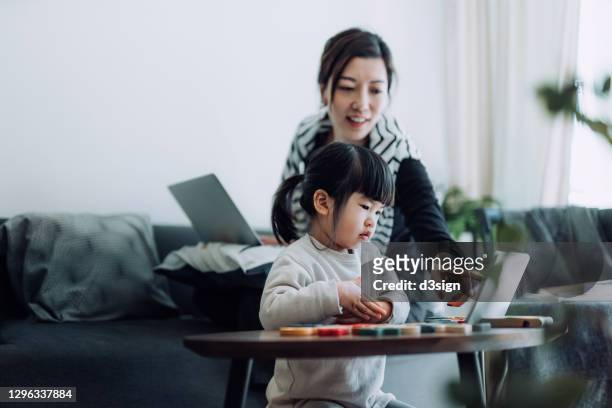 young asian mother working from home on laptop while homeschooling her daughter and assist her with school work. little daughter is studying from home and attending online school classes with a digital tablet. life in a quarantine - epidemie stock-fotos und bilder