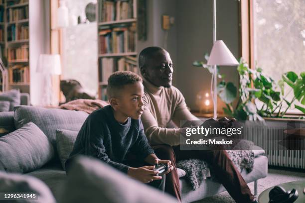 smiling father and son playing video game while sitting on sofa at home - son playing stock pictures, royalty-free photos & images