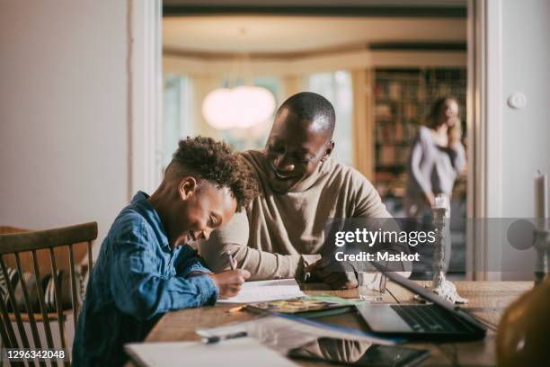 side view of smiling son writing while studying by father over table at home - ensino doméstico imagens e fotografias de stock