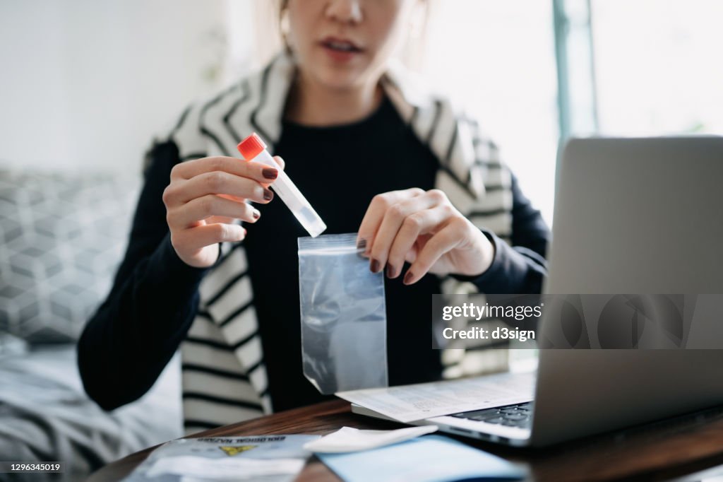 Cropped shot of young Asian woman consulting to her family doctor online in a virtual appointment, holding a medical test tube, conducting Covid-19 diagnostic test at home