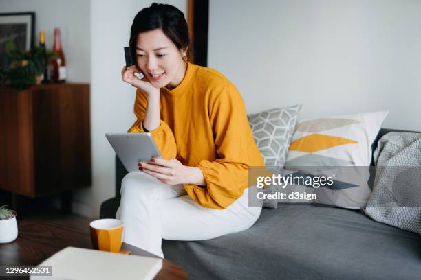 beautiful smiling young asian woman sitting on the sofa in the living room, shopping online with digital tablet and making mobile payment with credit card at cozy home. technology makes life so much easier - and booking com application stock pictures, royalty-free photos & images