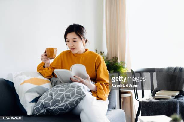 beautiful young asian woman reading a book while drinking a cup of coffee. enjoying a quiet time and relaxing environment at cozy home - reading foto e immagini stock