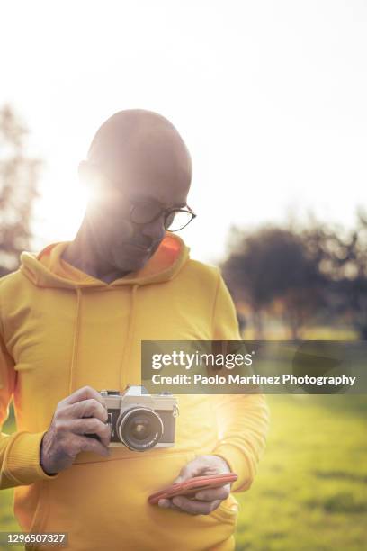 bald man holding analog camera while checking smartphone outdoor at sunset - examining lawn stock pictures, royalty-free photos & images