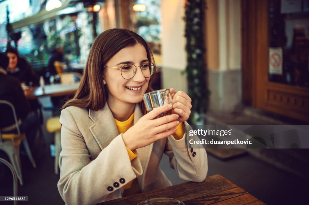 Smiling female influencer sitting on terrace of bar, holding cup of tea and warming up hands.